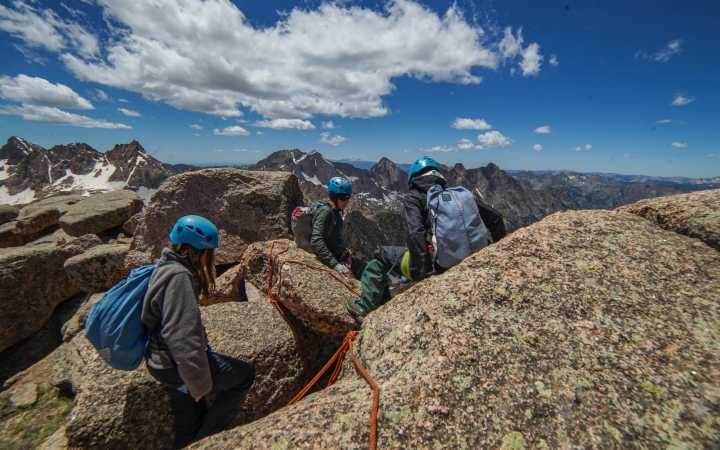 Three people wearing safety gear rest on rocks high above a mountainous landscape.